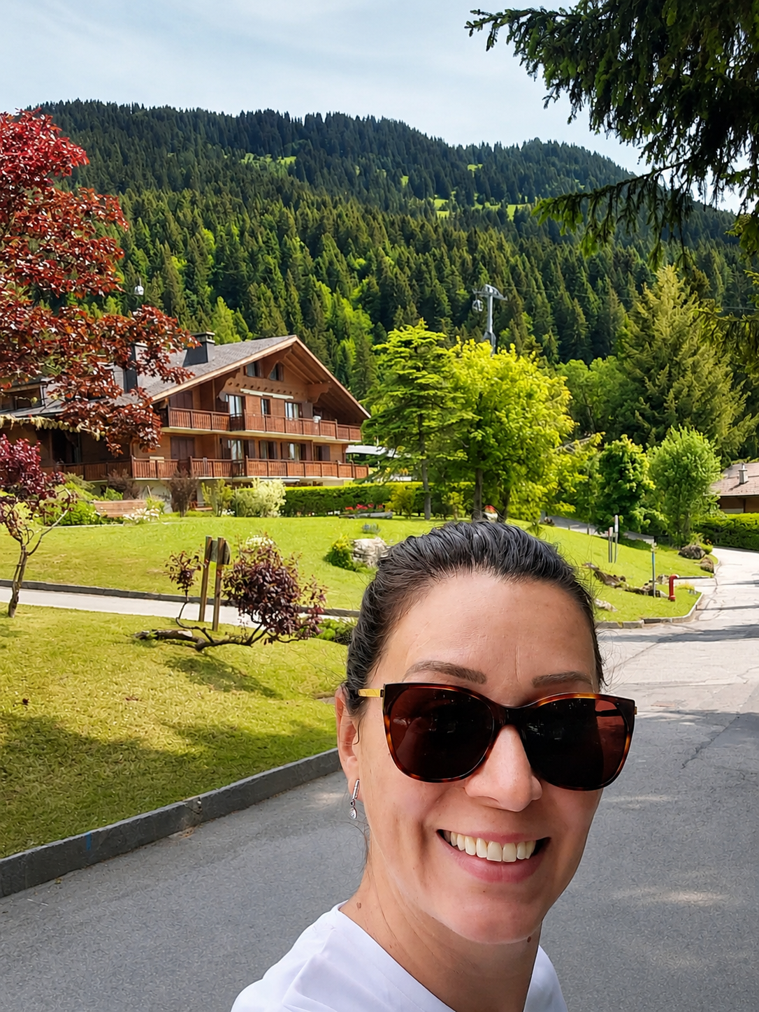 Giseli sorrindo nos Alpes suíços, com um chalé de madeira e a montanha ao fundo